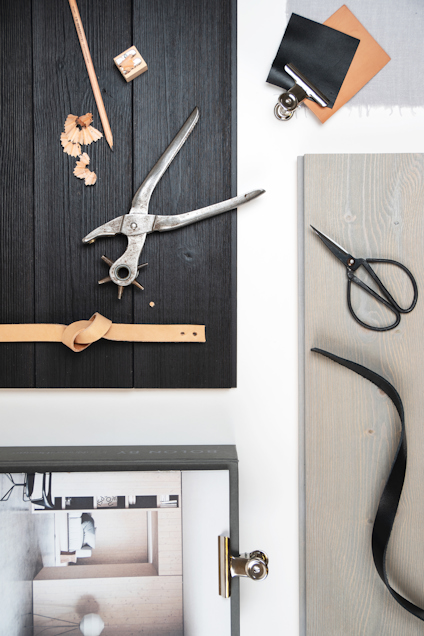 Svart träplanka med verktyg och träbitar, beige läderrem, sax på grå planka, papper och tygprover.