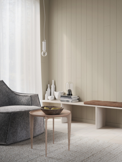 Ett modernt vardagsrum med grå fåtölj, rund träbänk, liten träbord med skål och hängande lampa framför en vit gardin.