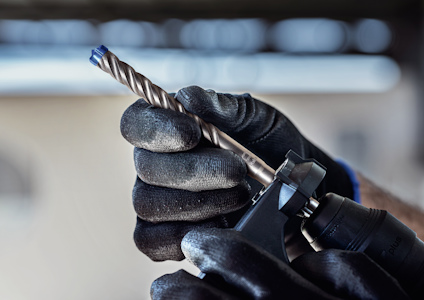 Svart handske håller en spiralborr med blå spets. Borrmaskin synlig.