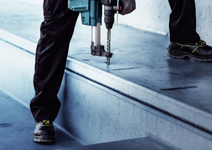 Person med borrmaskin borrar i betonggolv, klädd i skyddsskor och arbetsbyxor.