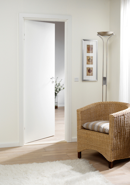 Öppen vit dörr, beige väggar, fåtölj av rotting, golvlampa, tre tavlor på väggen, ljus matta.