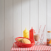 Vit träpanel bakom röd korg med bröd, ketchup- och senapsflaskor, glas, randiga sugrör och en flaska läsk på rutig duk.
