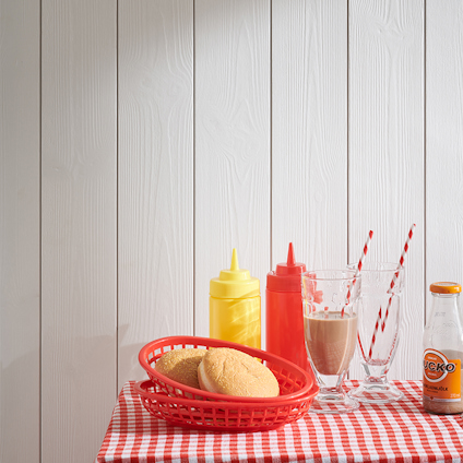 Vit träpanel bakom röd korg med bröd, ketchup- och senapsflaskor, glas, randiga sugrör och en flaska läsk på rutig duk.