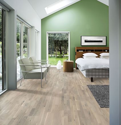 Sovrum med grön vägg, säng, grå fåtöljer, fönster med gardiner och ljus träparkett.