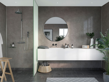 Modernt badrum med rund spegel, vit flytande diskbänk och dusch. Grön växt och trärstol kompletterar inredningen.