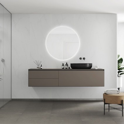 Modernt badrum med rund spegel, upplyst kant, och grå hängande förvaring. Svart handfat ovanpå skåpen.