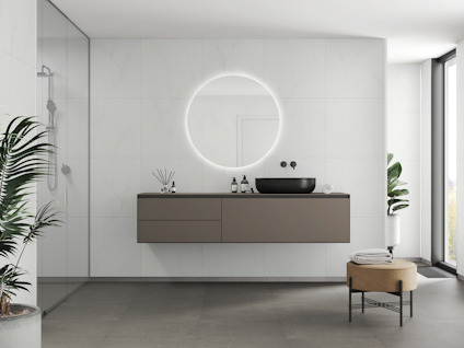 Modernt badrum med väggmonterat handfat, rund spegel med bakgrundsbelysning och dusch i glas. Grön växt i hörnet.