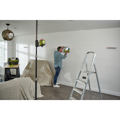 En person monterar hyllor på en vit vägg med hjälp av en grön borrmaskin och lasernivå. Stege i förgrunden.