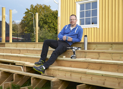 En person sitter på en trätrappa utomhus, med en termos och verktyg bredvid sig.