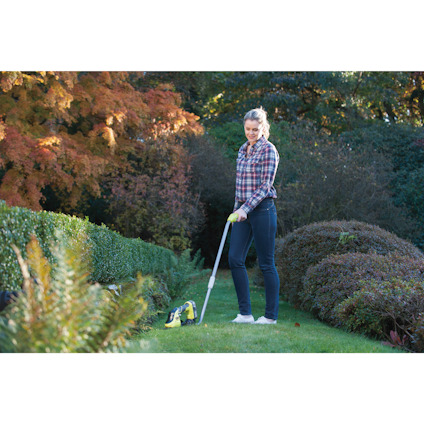 Person i rutiga skjorta och jeans använder trimmer i en trädgård med buskar och höstfärgade träd.