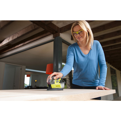 Kvinna använder en grön och svart slipmaskin på en trähylla i ett modernt hem. Hon bär skyddsglasögon.