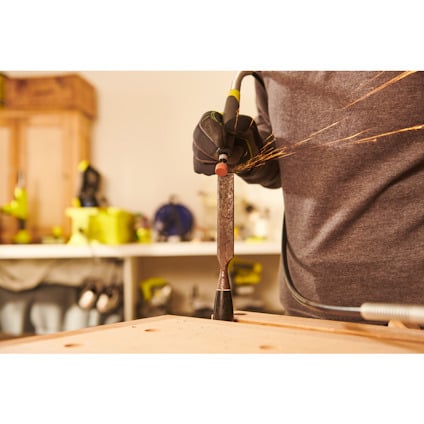 Person i arbetshandskar slipar mejsel med vinkelslip; gnistor flyger. Bakgrund: verkstadsutrustning.