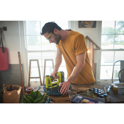 Man wearing a yellow shirt using a green nail gun on a workbench indoors.