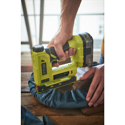 Person håller en grön batteridriven RYOBI-häftpistol över ett blått tyg på ett träbord.