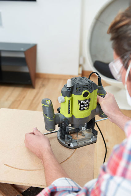 En person använder en grön och svart Ryobi-router för att bearbeta en träskiva.