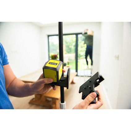 Person håller en laserverktygsmätare fastsatt på ett stativ inomhus, suddig bakgrund.