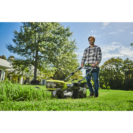 Man klipper gräs med en grön gräsklippare på en solig dag. Hus och träd i bakgrunden.