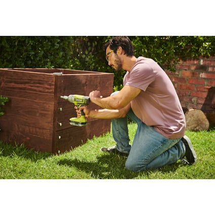 Person använder en grön och svart borrmaskin för att bygga en trästruktur på gräs.