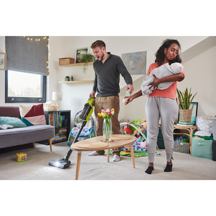 En person dammsuger medan en annan håller ett spädbarn i ett vardagsrum fyllt med leksaker och diverse föremål.