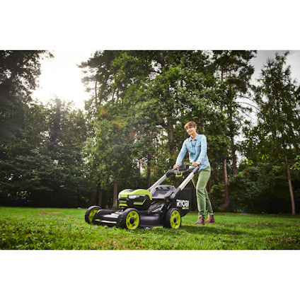 En person klipper gräs med en grön och svart gräsklippare från Ryobi på en solbelyst gräsmatta.