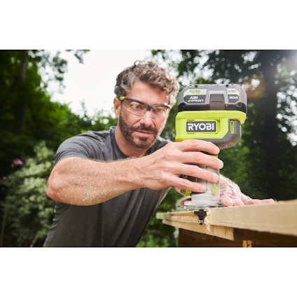En person använder en grön Ryobi sticksåg för att såga i trä utomhus, bär skyddsglasögon.