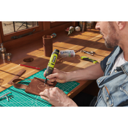 Person i jeansjacka använder en grön batteridriven borrmaskin på ett brunt läderstycke på arbetsbord.