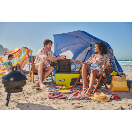 En man och en kvinna sitter på en sandstrand, omgivna av färgglada parasoller och en grill nära en RYOBI kylbox.