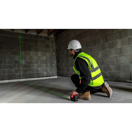 Person med hjälm och reflexväst använder en laserlinjeverktyg på ett betonggolv i ett ofärdigt rum.