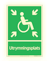Grön skylt med rullstolssymbol och fyra pilar pekande mot mitten. Texten "Utrymningsplats" längst ner.
