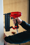 Person använder en röd och svart Senco tryckluftsspikpistol mot en vit vägg.