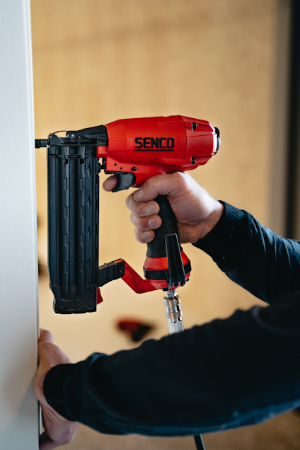 Person använder en röd och svart Senco tryckluftsspikpistol mot en vit vägg.