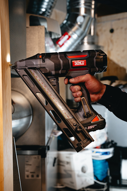 Person som håller i en spikpistol märkt "Senco" mot en träbit, med rör och industriell miljö i bakgrunden.