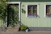 Grönt hus med två fönster med blomsterlådor, grön dörr och trappa. Växter bredvid dörren, vattenkanna på marken.