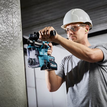 Person med vit hjälm använder en borrmaskin på en metallstruktur inomhus.