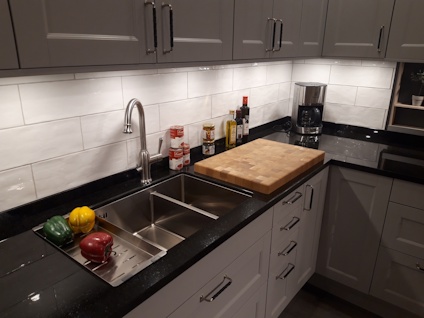 Modern kök med grå skåp, rostfri diskho, skärbräda, paprika och oljeflaskor på bänken. Kaklad vägg och bänkskiva i granit.