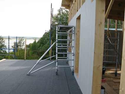 Byggställning vid husvägg, träd och vatten i bakgrunden.