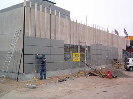 Byggarbetsplats med arbetare nära en fasad under uppförande, omgiven av stängsel och byggmaterial.