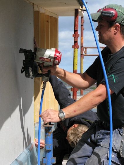Person använder en spikpistol på ett träpanel medan en annan person assisterar.