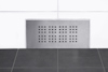 Rektangulärt ventilsystem i metall på en vit kakelvägg ovanför ett svart kakelgolv.