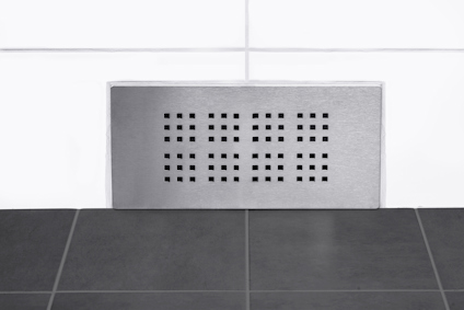 Rektangulärt ventilsystem i metall på en vit kakelvägg ovanför ett svart kakelgolv.