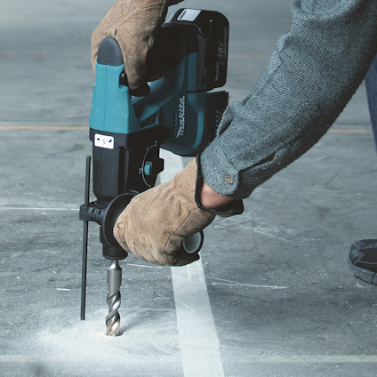En person borrar i ett betonggolv med en blå Makita borrmaskin, iklädd skyddshandskar.