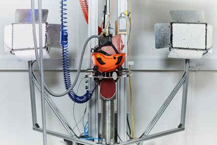 Robotarm med orange hjälm och slangar, omgiven av ljusreflektorer, i industriell miljö.