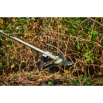 Gräs och slytrimmer av märket Ryobi i bruk skär igenom en tät vegetation av buskar och gräs.
