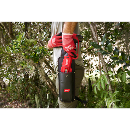Person wearing red gloves holding a tool in a black holster in a garden setting.