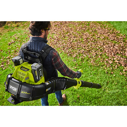 Person som använder en grön lövblås av märket Ryobi, bär en ryggsäck med extra batterier och står på gräs nära nedfallna löv