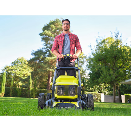 Man klipper gräsmattan med en grön och svart gräsklippare av märket Ryobi  i en trädgård.
