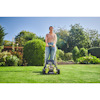 Kvinna använder en grön gräsklippare av märket Ryobi på en gräsmatta i en trädgård med buskar och träd i bakgrunden.