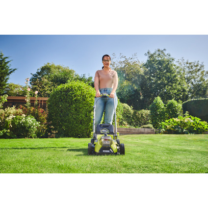 Kvinna använder en grön gräsklippare av märket Ryobi på en gräsmatta i en trädgård med buskar och träd i bakgrunden.