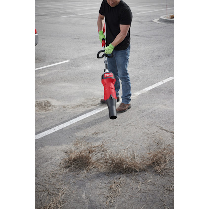 Person using a red leaf blower to clear dry grass from a parking lot.