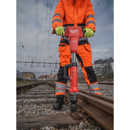 Person i orange arbetskläder använder en röd jackhammer nära järnvägsspår.